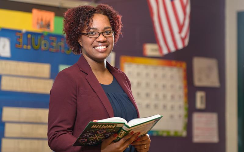 smiling teacher stands in classroom holding an English textbook