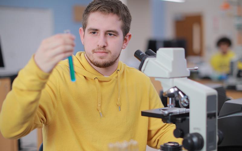 student in a classroom sitting infront of a microscope holding up a test tube full of liquid