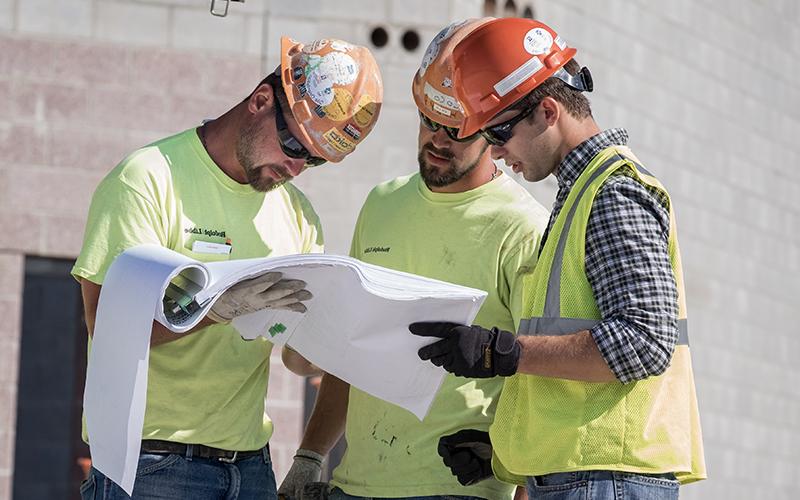 three men wearing orange hard hats and neon vests observe a page out of a large note pad file