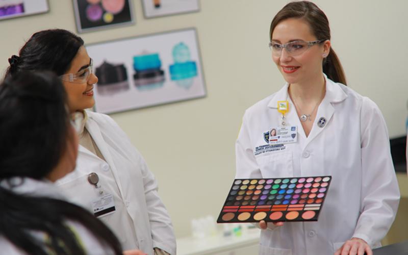 student in lab coat and goggles presents makeup palette to two other classmates