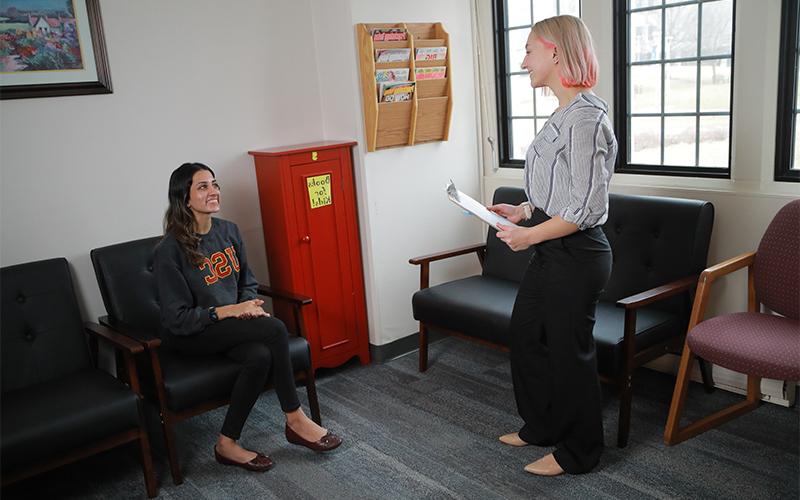 a woman standing holding a clipboard smiles and greets a woman sitting in a chair in a waiting room 