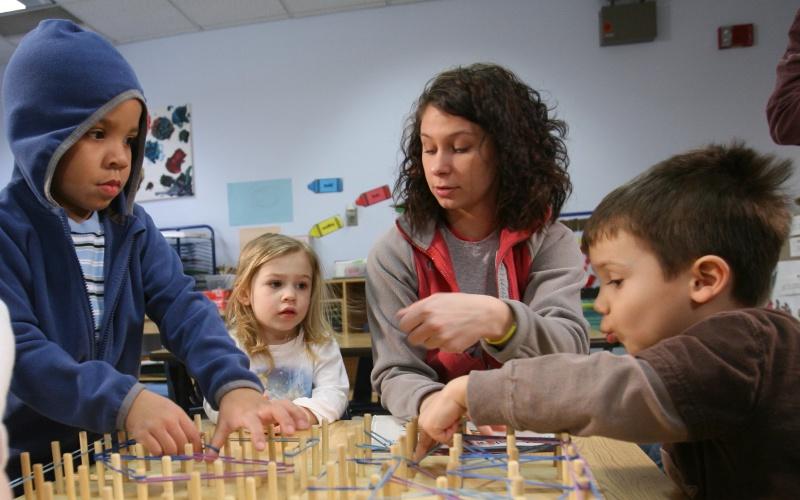 a teacher sits at a table with three young students during an activity of looping rubber bands onto a board with small wooden poles
