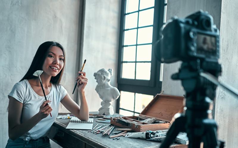 woman sits in front of camera and films video talking while holding two different paint brushes
