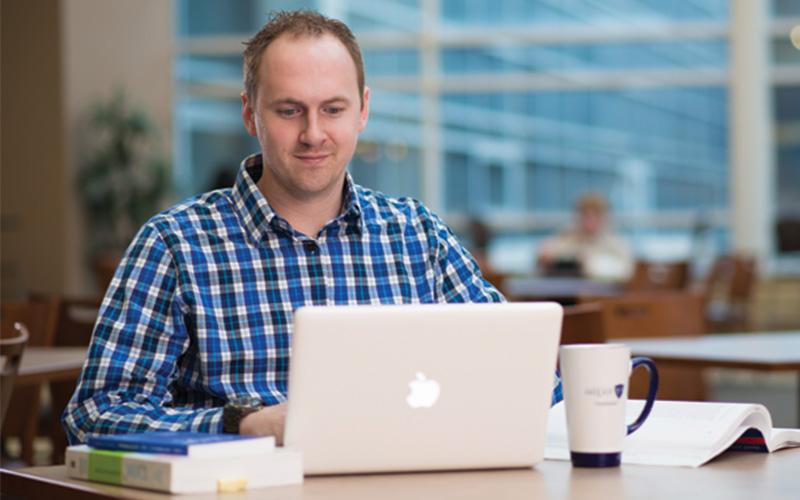 man sitting at table with open laptop and textbook