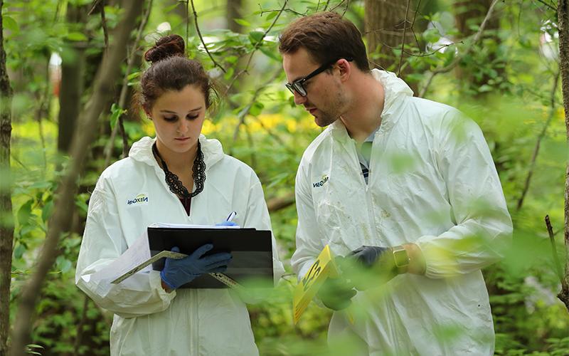 Male and female wearing safty equipment standing in the woods while the female writes on a clip board