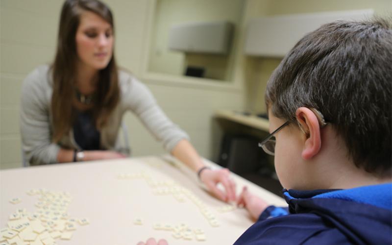 a woman sitting across a young child at a table helps piece together words using small tiles with letters 