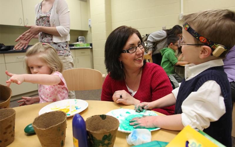 a woman crouches down at a table with two young children using paint for a craft