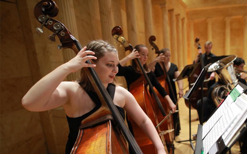 line of cello players perform from a music piece in a concert hall
