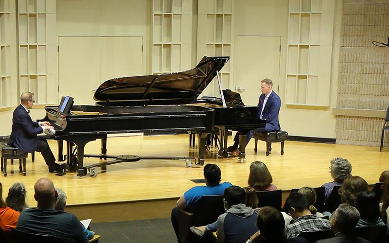 two men on piano perform on stage in a concert hall in front of an audience