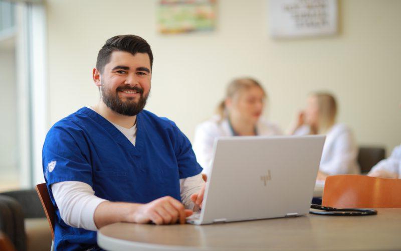 a man wering blue nursing scrubs sits at a table with an open laptop