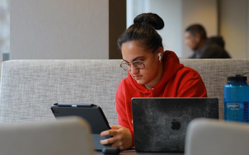 student studies in the library at a table, sitting in front of an open laptop and tablet