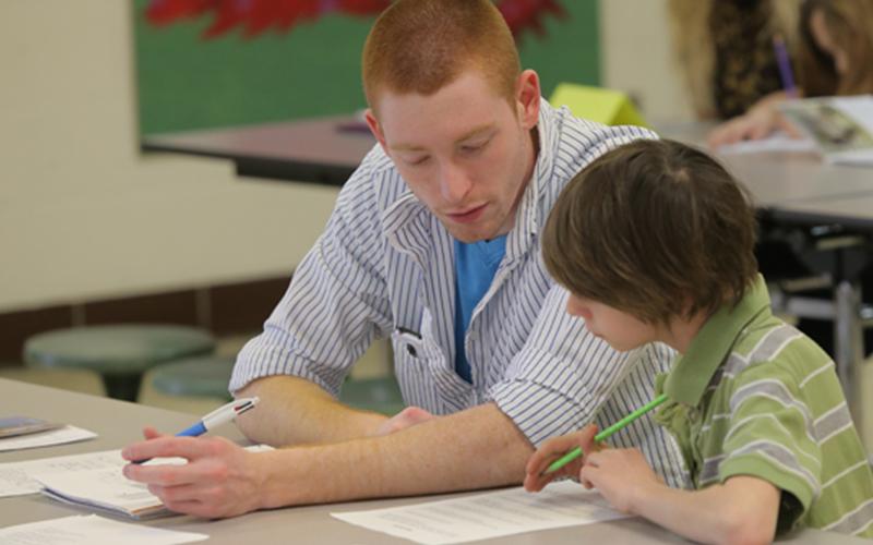 a man sits besides a child at a table in a classroom as the both look over one of the worksheets