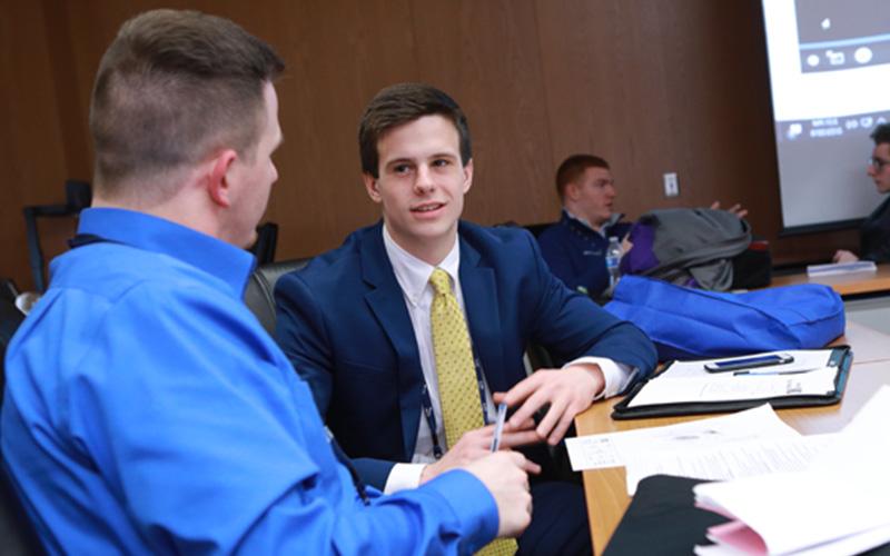 two sales students dressed in suits sit at a table to talk with notes open 