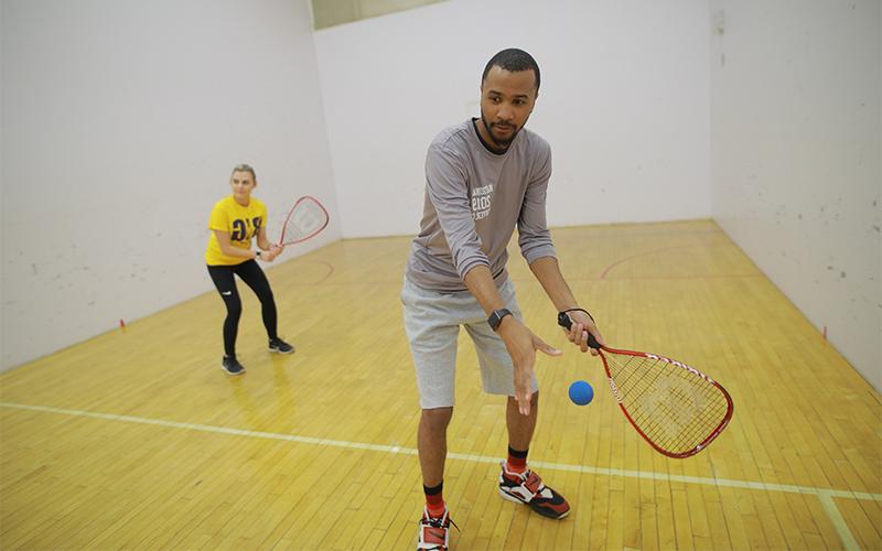 a man and woman in an indoor racquetball court prepare to serve the ball