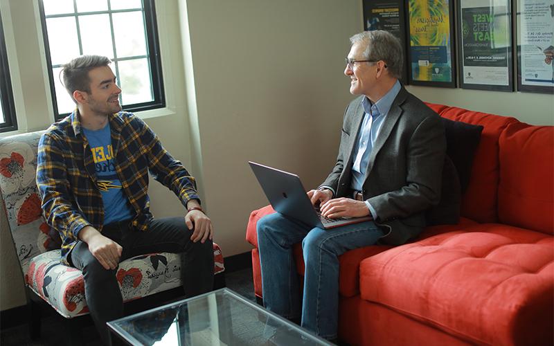 a man sitting on a couch with an open laptop on his lap talks to the man seated adjacently in the chair next to him