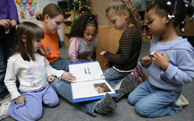 a woman sits on the floor with a small group of young children and reads from a blue binder