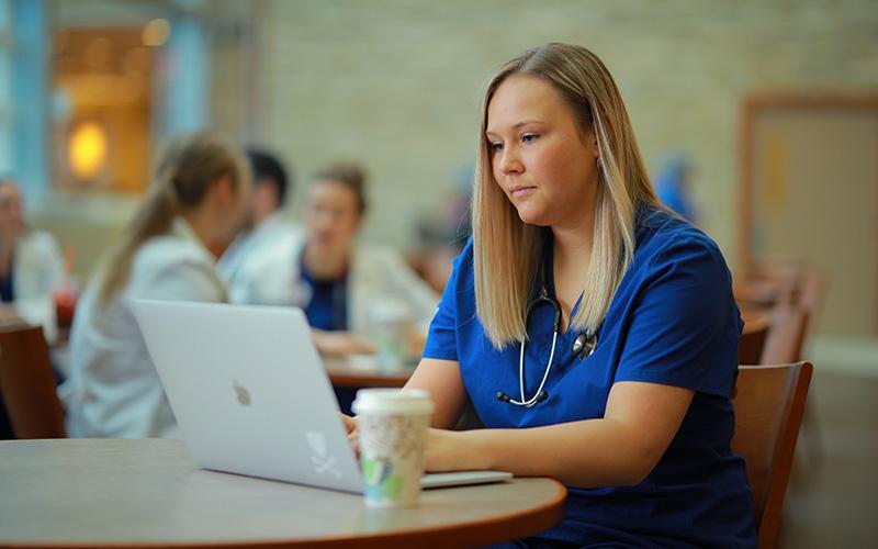 student in blue scrubs sits at table in front of an open laptop